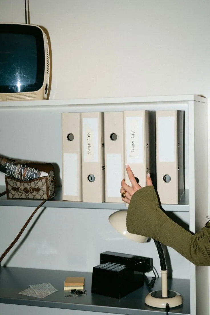 A hand reaching for one of a row of labelled box files on a shelf above a desk